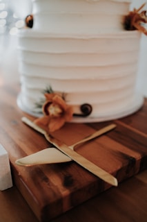 A close-up of a tiered white cake with simple, elegant decorations. The cake has a textured finish and is adorned with small floral arrangements featuring orange flowers and other natural elements. It is situated on a wooden board with a gold cake knife and server beside it.