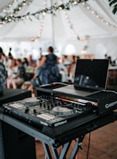 A DJ setup with a laptop and mixing console is positioned prominently in the foreground, set up on a stand. Behind it, a blurred background shows people sitting and standing under a large white tent adorned with string lights and greenery, suggesting an event or celebration.