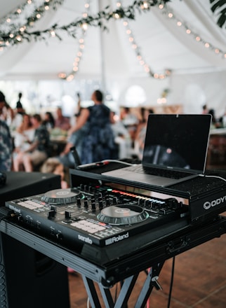 A DJ setup with a laptop and mixing console is positioned prominently in the foreground, set up on a stand. Behind it, a blurred background shows people sitting and standing under a large white tent adorned with string lights and greenery, suggesting an event or celebration.