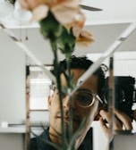 A person is taking a photograph using a round camera through a mirror that is segmented into multiple triangles. There is a blurred image of a plant with light pink flowers in the foreground, which creates an artistic and somewhat obscure visual effect.