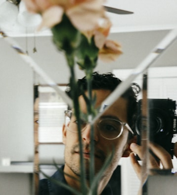 A person is taking a photograph using a round camera through a mirror that is segmented into multiple triangles. There is a blurred image of a plant with light pink flowers in the foreground, which creates an artistic and somewhat obscure visual effect.