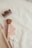 Items such as a hair claw clip, a makeup brush with a wooden handle, a compact mirror with a pink cover, and a wrapped tampon are arranged on a cream-colored fabric surface. These objects are partially tucked into a beige, textured zip pouch.