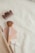 Items such as a hair claw clip, a makeup brush with a wooden handle, a compact mirror with a pink cover, and a wrapped tampon are arranged on a cream-colored fabric surface. These objects are partially tucked into a beige, textured zip pouch.