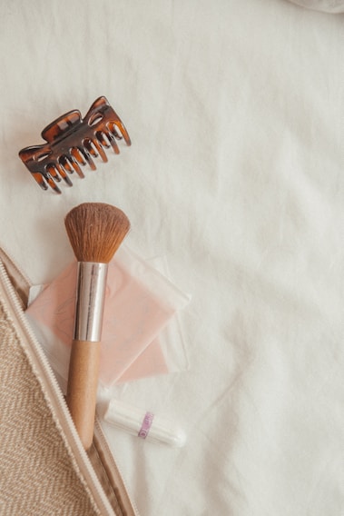 Items such as a hair claw clip, a makeup brush with a wooden handle, a compact mirror with a pink cover, and a wrapped tampon are arranged on a cream-colored fabric surface. These objects are partially tucked into a beige, textured zip pouch.
