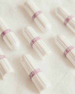 Several white tampons arranged on a soft, white fabric surface. Each tampon is wrapped in individual packaging with a purple band labeled 'TOM'. The arrangement is neat and evenly spaced.