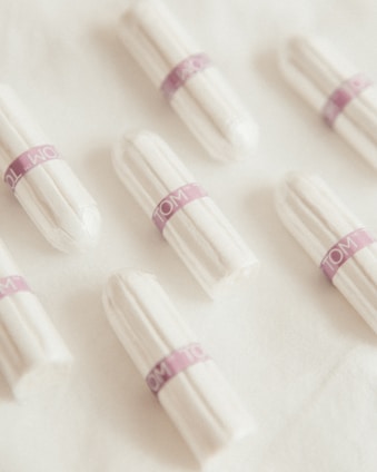 Several white tampons arranged on a soft, white fabric surface. Each tampon is wrapped in individual packaging with a purple band labeled 'TOM'. The arrangement is neat and evenly spaced.