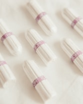 Several white tampons arranged on a soft, white fabric surface. Each tampon is wrapped in individual packaging with a purple band labeled 'TOM'. The arrangement is neat and evenly spaced.