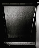 A dirty window is slightly ajar, allowing a view of a dark exterior. The window glass is covered with water stains or grime, and there is a tiled window sill visible at the bottom. The overall scene appears neglected and dimly lit.