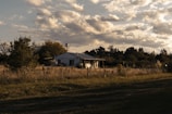 A rural house is situated in an open field, surrounded by a wire fence and various trees. The sky is partly cloudy, with sunlight casting warm tones over the landscape. The house appears small and simple, blending into the natural scenery.
