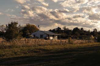 A rural house is situated in an open field, surrounded by a wire fence and various trees. The sky is partly cloudy, with sunlight casting warm tones over the landscape. The house appears small and simple, blending into the natural scenery.
