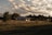 A rural house is situated in an open field, surrounded by a wire fence and various trees. The sky is partly cloudy, with sunlight casting warm tones over the landscape. The house appears small and simple, blending into the natural scenery.