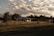 A rural house is situated in an open field, surrounded by a wire fence and various trees. The sky is partly cloudy, with sunlight casting warm tones over the landscape. The house appears small and simple, blending into the natural scenery.