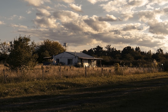 A rural house is situated in an open field, surrounded by a wire fence and various trees. The sky is partly cloudy, with sunlight casting warm tones over the landscape. The house appears small and simple, blending into the natural scenery.