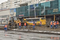 Workers in blue and orange uniforms are engaged in construction activity on a city street. A bulldozer is parked nearby, and a yellow bus is visible in the background. The scene includes several buildings with signage in a foreign language.