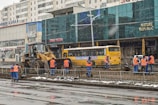 Workers in blue and orange uniforms are engaged in construction activity on a city street. A bulldozer is parked nearby, and a yellow bus is visible in the background. The scene includes several buildings with signage in a foreign language.