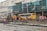 Workers in blue and orange uniforms are engaged in construction activity on a city street. A bulldozer is parked nearby, and a yellow bus is visible in the background. The scene includes several buildings with signage in a foreign language.