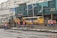 Workers in blue and orange uniforms are engaged in construction activity on a city street. A bulldozer is parked nearby, and a yellow bus is visible in the background. The scene includes several buildings with signage in a foreign language.