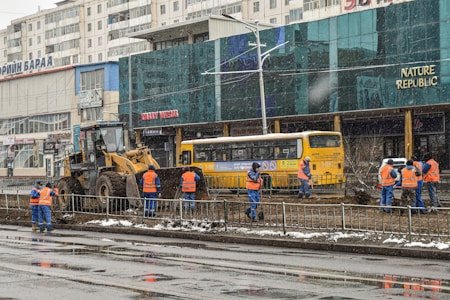 Workers in blue and orange uniforms are engaged in construction activity on a city street. A bulldozer is parked nearby, and a yellow bus is visible in the background. The scene includes several buildings with signage in a foreign language.