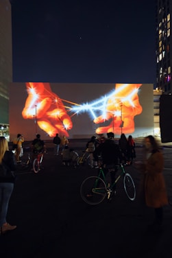 A nighttime scene features a large building facade with a vivid digital artwork projected onto it, showing two illuminated hands interacting with a blue, sparkling line that resembles electricity or energy. Below the projection, a group of people is gathered, some standing and others seated on bicycles, observing the display. Streetlights and buildings with illuminated windows frame the scene, adding to the urban atmosphere.