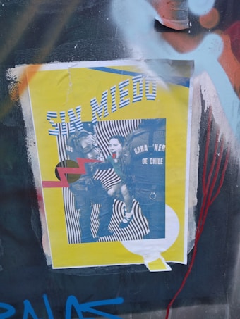 A poster is pasted on a surface with graffiti. The poster has a yellow background with black and white images in the center, depicting a scene involving law enforcement officers and a person being restrained. One part of the text reads 'CARABINEROS DE CHILE', and there is large text at the top saying 'SIN MIEDO'. Red and black graphic elements, including zigzag lines, overlay the image.