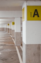 A series of concrete pillars in a parking garage are marked with bright yellow signage indicating Zone A. The floor is clean and lightly marked with white lines, creating a sense of order.