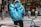 A delivery person wearing a blue jacket and a large blue backpack with the logo 'stuart Same Day Delivery' is standing next to a blue bicycle. The person is facing away from the camera, and they are near storefronts including a Zara store. Other people are visible in the background, along with various street elements such as bollards and signs.