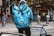 A delivery person wearing a blue jacket and a large blue backpack with the logo 'stuart Same Day Delivery' is standing next to a blue bicycle. The person is facing away from the camera, and they are near storefronts including a Zara store. Other people are visible in the background, along with various street elements such as bollards and signs.
