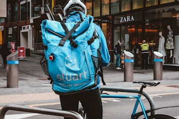 A delivery person wearing a blue jacket and a large blue backpack with the logo 'stuart Same Day Delivery' is standing next to a blue bicycle. The person is facing away from the camera, and they are near storefronts including a Zara store. Other people are visible in the background, along with various street elements such as bollards and signs.