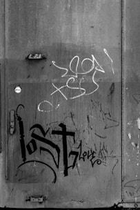 A gritty urban wall features various graffiti tags and marks, including the word 'lost' prominently displayed in bold, stylized letters. The wall is weathered and textured, with remnants of peeled paint and signs of wear, contributing to an overall atmosphere of neglect or decay.