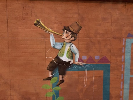 A mural depicts a cartoonish figure sitting on a ledge, blowing a long horn. The figure wears a vest, trousers, and a hat, with exaggerated features characteristic of street art. A geometric pattern decorates the wall, and a green vine grows from below.