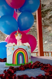 A colorful cake decorated with a fondant rainbow and clouds, topped with a unicorn figurine, surrounded by vibrant blue and pink balloons. The cake features stars and is placed on a table with a dark cloth and red petals.