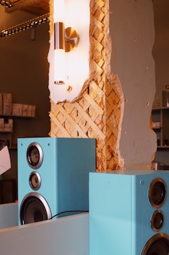 A partially deconstructed wall reveals its wooden lattice interior next to a sleek modern wall-mounted light fixture. Large blue speakers are placed in the foreground, adding a pop of color and contrast to the rustic texture of the exposed wall.