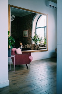 A stylish interior featuring a large wall mirror reflecting a cozy seating area with patterned cushions and potted plants. The space has a rustic brick wall contrasted by a contemporary glass window. A pink armchair and a small side table are in the foreground with wooden flooring throughout.