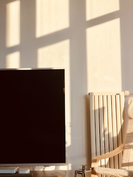 Sunlight casts long rectangular shadows and light patterns on a plain white wall, partially illuminating a flat screen television positioned on a table. To the right of the television, a beige radiator is present, alongside a light-colored wooden chair with a fabric cushion. The overall scene is bathed in warm sunlight, creating a serene atmosphere.