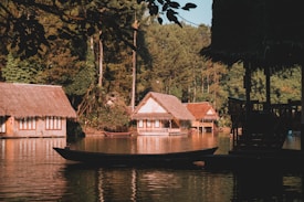 Several traditional thatched-roof cottages are situated along the edge of a calm body of water surrounded by lush green trees. A small wooden boat floats in the foreground, partially in shadow due to overhanging branches. The scene has a tranquil and serene ambiance, enhanced by the warm lighting from the setting sun.