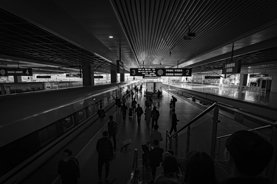 A black and white photo of a busy train station platform with people walking, some carrying luggage. The platform is covered by a large roof structure, with signs indicating platform numbers. A long train is stationed on the left, and the area is well-lit with a skylight visible above.