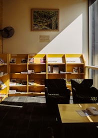 A cozy reading area with two black armchairs positioned beside a wooden table, situated in front of a set of yellow bookshelves stocked with various magazines and publications. Sunlight streams through a large window, casting sharp shadows on the floor and partially illuminating the shelves. A framed painting and a wall-mounted notice are displayed on the white-painted wall above the bookshelves.