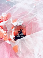 A perfume bottle with intricate design details, placed against a backdrop of soft pink and white mesh fabric. Surrounding the bottle are scattered heart-shaped cutouts in red and white.