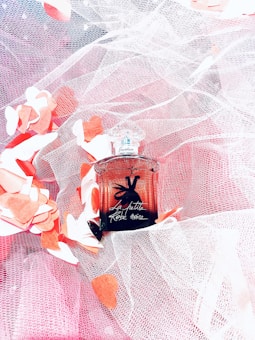 A perfume bottle with an elegant design labeled 'La Petite Robe Noire' rests on a bed of white and pink lace fabric. Surrounding the bottle are scattered paper hearts in shades of red and white.