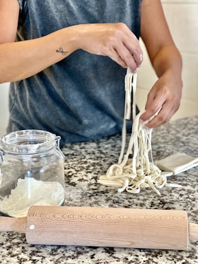 A person is making noodles by hand, with a clear view of a rolling pin, a glass jar of flour, and a bundle of freshly made noodles on a speckled granite countertop. The individual's arms are visible, along with a specific tattoo on one arm. The setting appears to be a kitchen.