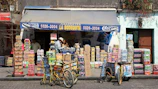 A small corner store with a blue awning labeled with 'La Bodeguita' and various products including drinks and snacks stacked in front. Two people are seen interacting near the entrance, and two bicycles with baskets are parked nearby. The facade of the building is weathered, suggesting an urban, lively environment.