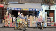 A small corner store with a blue awning labeled with 'La Bodeguita' and various products including drinks and snacks stacked in front. Two people are seen interacting near the entrance, and two bicycles with baskets are parked nearby. The facade of the building is weathered, suggesting an urban, lively environment.