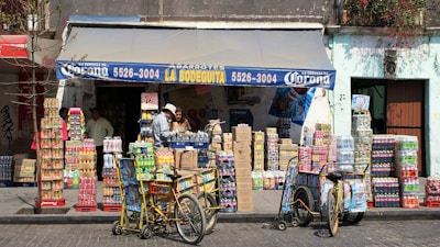 A small corner store with a blue awning labeled with 'La Bodeguita' and various products including drinks and snacks stacked in front. Two people are seen interacting near the entrance, and two bicycles with baskets are parked nearby. The facade of the building is weathered, suggesting an urban, lively environment.