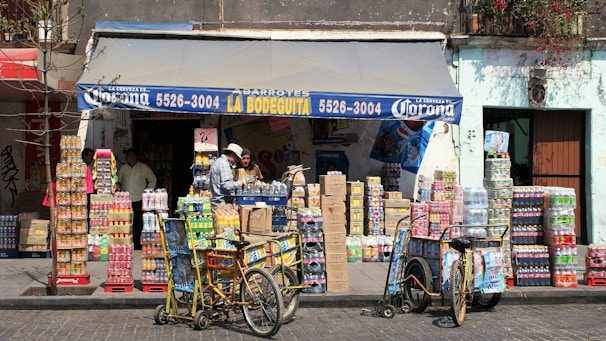 A small corner store with a blue awning labeled with 'La Bodeguita' and various products including drinks and snacks stacked in front. Two people are seen interacting near the entrance, and two bicycles with baskets are parked nearby. The facade of the building is weathered, suggesting an urban, lively environment.