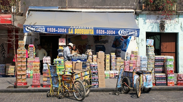 A small corner store with a blue awning labeled with 'La Bodeguita' and various products including drinks and snacks stacked in front. Two people are seen interacting near the entrance, and two bicycles with baskets are parked nearby. The facade of the building is weathered, suggesting an urban, lively environment.