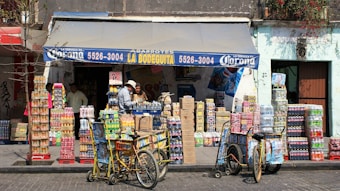 A small corner store with a blue awning labeled with 'La Bodeguita' and various products including drinks and snacks stacked in front. Two people are seen interacting near the entrance, and two bicycles with baskets are parked nearby. The facade of the building is weathered, suggesting an urban, lively environment.