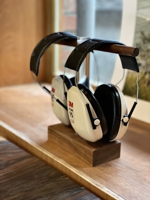 A pair of 3M earmuff hearing protectors with a noise reduction rating of 95, resting on a wooden stand placed on a wooden surface. The earmuffs have a black headband and white ear cups, with labels indicating the brand and model. A softly focused window and partial view of an outdoor scene are visible in the background.