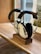 A pair of 3M earmuff hearing protectors with a noise reduction rating of 95, resting on a wooden stand placed on a wooden surface. The earmuffs have a black headband and white ear cups, with labels indicating the brand and model. A softly focused window and partial view of an outdoor scene are visible in the background.