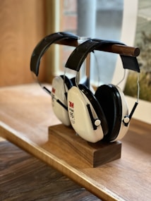 A pair of 3M earmuff hearing protectors with a noise reduction rating of 95, resting on a wooden stand placed on a wooden surface. The earmuffs have a black headband and white ear cups, with labels indicating the brand and model. A softly focused window and partial view of an outdoor scene are visible in the background.