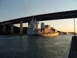 A large cargo ship carrying numerous containers sails under a tall bridge spanning a calm waterway. The lighting suggests late afternoon or early evening, with the sky showing tones of orange and blue. On the horizon, there are industrial buildings and trees along the riverbank.